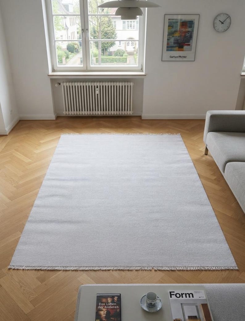 May include: A rectangular light gray area rug with a flat-woven texture and short fringe detailing along the top and bottom edges. The rug is positioned centrally on a light brown wooden floor with a herringbone pattern, featuring a solid color.