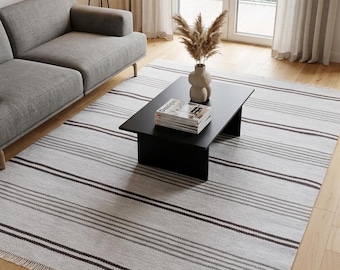Puede incluir: Una alfombra rectangular con una base de color gris claro y rayas horizontales en varios tonos de gris oscuro y negro. La alfombra presenta un tejido texturizado y flecos cortos en los dos bordes más cortos, ofreciendo un diseño lineal y limpio.