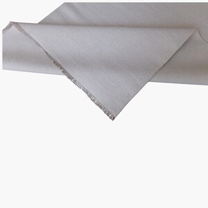 May include: A piece of light gray fabric with a visible woven texture. The material is folded over at an angle, revealing a finished edge with a silver-colored metallic trim that runs along the length of the seam against the plain fabric.