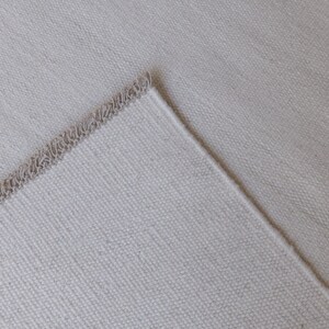 May include: A light beige fabric with a visible woven texture and a finished edge featuring a decorative, looped fringe detail. The material has a consistent, neutral tone and a structured, durable appearance with a clear, repeating weave pattern.
