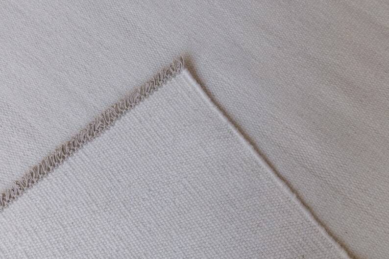 May include: A light beige fabric with a visible woven texture and a finished edge featuring a decorative, looped fringe detail. The material has a consistent, neutral tone and a structured, durable appearance with a clear, repeating weave pattern.