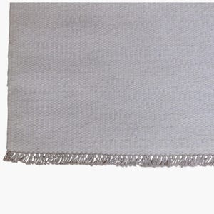 May include: A rectangular area rug featuring a light gray, woven textile surface with a consistent, dense, and textured pattern. The bottom edge of the rug is finished with a row of short, matching light gray fringe tassels along the entire width.