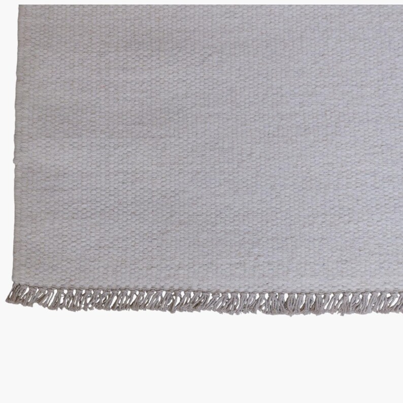 May include: A rectangular area rug featuring a light gray, woven textile surface with a consistent, dense, and textured pattern. The bottom edge of the rug is finished with a row of short, matching light gray fringe tassels along the entire width.