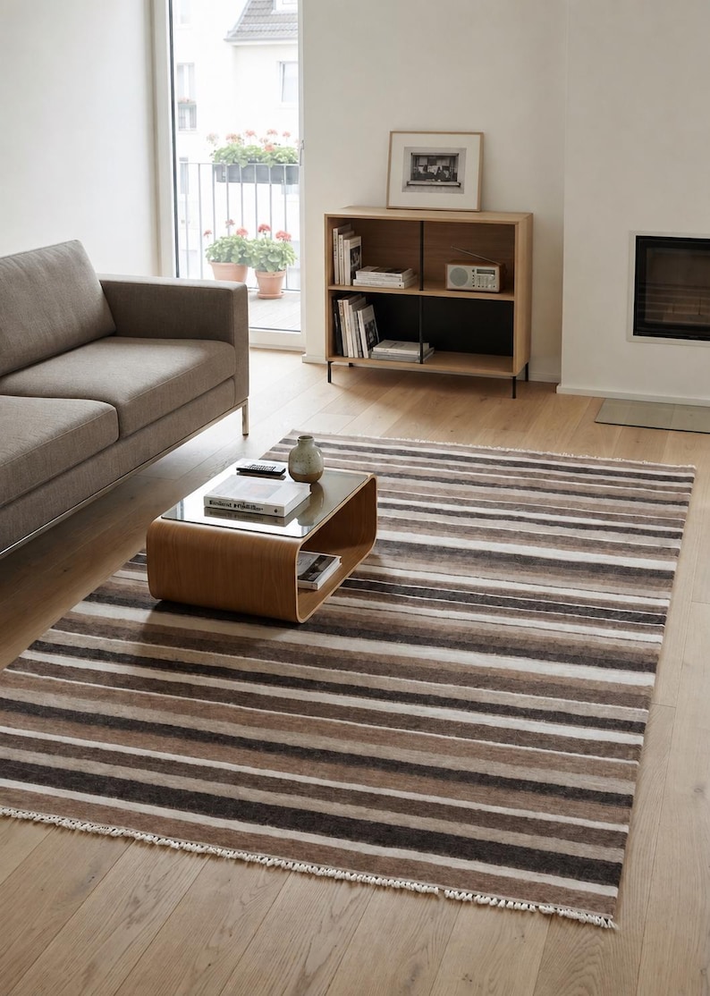 Puede incluir: Una alfombra rectangular con rayas horizontales en varios tonos de marr&oacute;n, beige, crema y gris oscuro. La alfombra tiene una apariencia suave y texturizada con flecos blancos cortos a lo largo de los dos bordes m&aacute;s cortos de la tela.