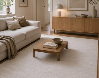 May include: A large, rectangular, solid off-white area rug with a soft, textured pile surface. The rug is positioned on a light-colored wooden floor, extending from beneath a light-colored sofa to the center of the room, providing a neutral foundation.
