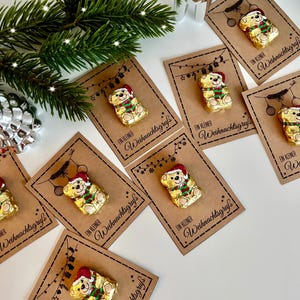 Può includere: Biglietti regalo natalizi con cioccolatini a forma di orsetto avvolti in carta dorata, con cappelli e sciarpe di Babbo Natale. I biglietti sono marroni con il testo tedesco "Ein kleiner Weihnachtsgruß" e luci decorative.