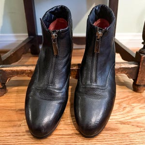 May include: A pair of black leather ankle boots with a front zipper closure. The boots have a rounded toe and a low heel. The interior of the boots is lined with a red material. The boots are displayed on a wooden surface.