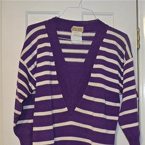 May include: A purple and white striped sweater with a deep V-neck. The sweater has three-quarter sleeves and is hanging on a white hanger. The label reads "Lina Lee".