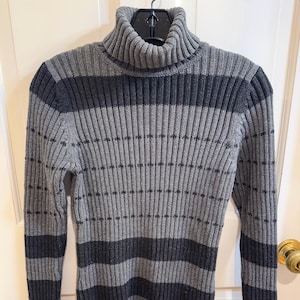 Puede incluir: Un jersey de cuello alto a rayas grises y gris oscuro. El jersey de punto acanalado tiene un cuello alto y doblado y mangas largas. Las rayas grises oscuras son horizontales y recorren el pecho y las mangas.