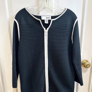 May include: Black zip-up sweater with white trim and a ribbed texture. The sweater has long sleeves and a white zipper. The brand name "Joan Vass" is visible on a tag near the neckline. The sweater is hanging on a clear hanger.