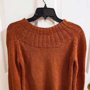 May include: A rust-colored sweater with a ribbed, high neckline and long sleeves. The sweater is hanging on a black hanger against a white door. The knitwear has a textured appearance.