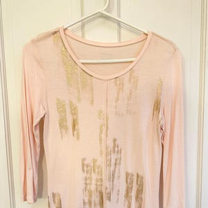 May include: A light pink long-sleeve shirt with a scoop neck and gold brushstroke design. The shirt is hanging on a white hanger against a white background.