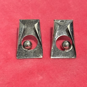 May include: A pair of silver-toned metal earrings featuring a trapezoidal frame with folded top corners and a circular cutout in the center. Each earring contains a small, polished silver sphere suspended within the circular opening.