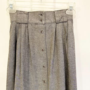 May include: A gray button-down skirt hangs on a clear plastic hanger. The skirt has a high waist with a wide waistband and pleats. It features a series of buttons down the front. The fabric appears to be a textured, woven material.