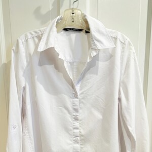 May include: A crisp white long-sleeve button-down shirt featuring a classic collar and a front button placket. The garment includes a label at the neckline with the text Zac & Rachel. The sleeves have a tab detail for rolling up the cuffs.