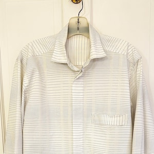 May include: A cream-colored long-sleeve shirt with a subtle striped pattern in dark gray. The shirt features a classic collar, a single chest pocket, and is hanging on a clear plastic hanger.