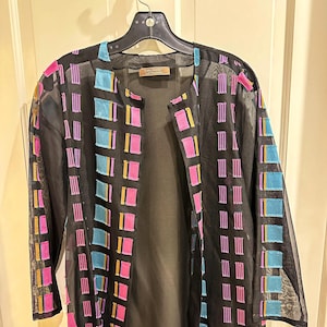May include: A black, open-front jacket with long sleeves. The jacket is adorned with a grid pattern of colorful squares and vertical stripes in shades of pink, blue, and yellow. The jacket is hanging on a black hanger.