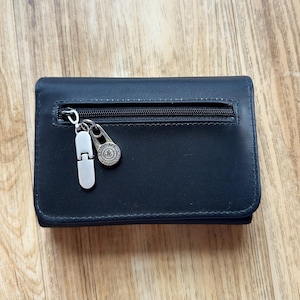 May include: A black leather wallet with a zippered pocket and a silver zipper pull. The wallet has a small silver charm attached to the zipper. The wallet is closed and sitting on a wooden surface.