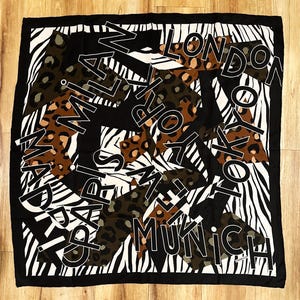 May include: Square black and white scarf with a bold design. The scarf features a zebra print, leopard print, and the names of cities like Paris, Milan, and London in white lettering. The scarf has a black border.