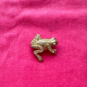 May include: A gold-toned frog brooch with clear crystal accents. The frog is detailed with textured skin and sparkling crystals on its body and legs. The brooch is set against a bright pink background.