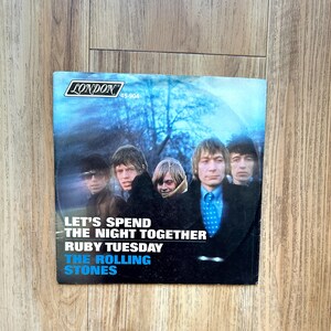 May include: A vintage vinyl record sleeve for The Rolling Stones featuring the songs Let's Spend The Night Together and Ruby Tuesday. The cover displays a group portrait of the band members against a blue-toned outdoor background with London branding.