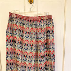 May include: A colorful skirt with a repeating pattern of red, blue, and green floral designs. The skirt has a red waistband and is fastened with gold-colored buttons. The skirt is hanging on a hanger against a white door.