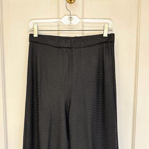 May include: Black, textured pants hanging on a clear plastic hanger. The pants have a subtle, repeating pattern and a wide waistband. The fabric appears to be lightweight.