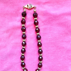 May include: A necklace featuring faceted, deep red beads interspersed with gold-colored spacers. The necklace is secured with a gold-toned clasp. The beads have a sparkling, jewel-like appearance, and the necklace is displayed against a pink background.