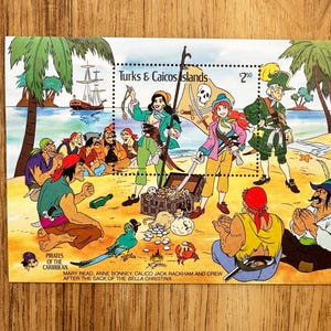 May include: A postage stamp sheet from the Turks & Caicos Islands featuring a colorful cartoon illustration of pirates on a beach. The scene includes a treasure chest, a skull and crossbones flag, a ship in the distance, and text identifying the figures as Mary Read, Anne Bonney, Calico Jack Rackham and crew after the sack of the Bella Christina.