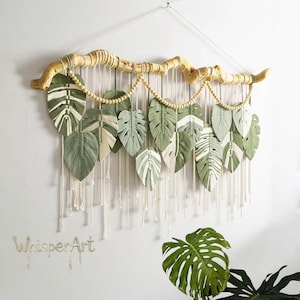 May include: A decorative macrame wall hanging with green and white painted leaf designs suspended from a natural wooden branch. The leaves are arranged in a cascading pattern, with wooden beads and hanging cords. The text "WhisperArt" is visible.