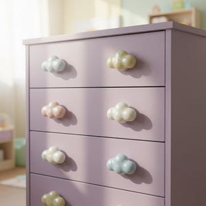 May include: A tall, light purple dresser featuring four horizontal drawers. Each drawer is equipped with two cloud-shaped knobs in various pastel colors, including soft blue, pale yellow, and light pink, creating a whimsical and organized storage unit.