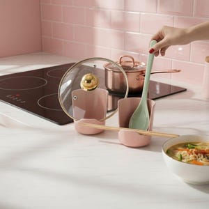 Puede incluir: Dos soportes para utensilios de cocina de color rosa claro diseñados para sostener una tapa de olla y una cuchara. El conjunto incluye una cuchara de color verde claro y un par de palillos de madera apoyados sobre los soportes en una encimera.