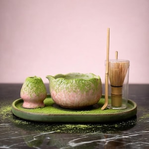Puede incluir: Un juego de té matcha sobre una bandeja verde. El juego incluye un cuenco rosa y verde, un batidor, una cuchara y un recipiente pequeño. El polvo de matcha es verde y cubre el cuenco y el recipiente pequeño. El fondo es rosa.