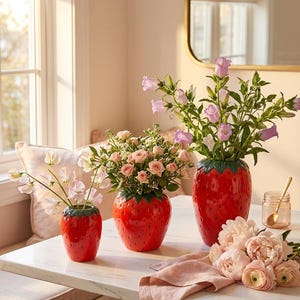 以下が含まれることがあります： イチゴの形をした光沢のある赤い陶器製の花瓶3つ。緑色の葉の縁取りが施されています。花瓶はサイズが異なり、それぞれ淡いピンクのスイートピー、小さなピンクのバラ、紫色のベルフラワーなど、異なるフラワーアレンジメントが飾られています。