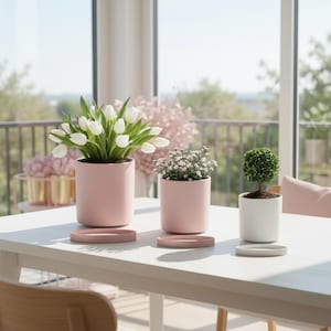 以下が含まれることがあります： 白いテーブルの上に3つの鉢植え。2つのピンク色の円筒形の鉢には白いチューリップと小さな白い花が入っています。白い鉢には小さくて丸い緑色の植物が入っています。各鉢にはお揃いの受け皿が付いています。