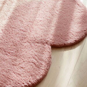 Peut inclure: Gros plan sur un tapis en peluche rose doux de forme arrondie. Le tapis a une surface textur&eacute;e et est pos&eacute; sur un plancher en bois clair. La couleur du tapis est un rose poudr&eacute;.