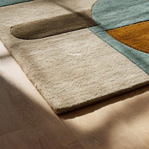 May include: A close-up of a modern area rug with a geometric design. The rug features a beige background with brown, teal, and orange shapes. The rug is made of a soft, textured material.