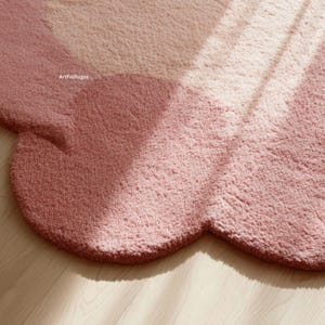 Peut inclure: Tapis en forme de nuage dans des tons roses. Le tapis pr&eacute;sente un bord festonn&eacute; et une surface textur&eacute;e. Le nom de la marque "ArtfulRugss" est visible. Le tapis est pos&eacute; sur un sol en bois clair.