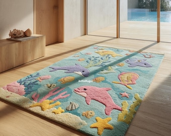 Op de afbeelding: Een rechthoekig vloerkleed met een lichtblauwe achtergrond en een getuft onderwatertafereel. Het ontwerp bevat een roze dolfijn, een paarse dolfijn, gele zeesterren, koraalformaties, diverse kleine vissen en schelpen. De tekst ArtfulRugss is zichtbaar.