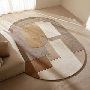May include: An oval-shaped area rug with a geometric design in shades of beige, brown, and cream. The rug features a mix of rectangular and curved shapes, creating a modern and abstract pattern. The rug is placed on a light-colored wooden floor.