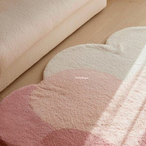 Peut inclure: Un tapis en forme de nuage aux tons rose et cr&egrave;me. Le tapis a un aspect doux et textur&eacute; et est pos&eacute; sur un sol en bois clair. Le design pr&eacute;sente un d&eacute;grad&eacute; de couleurs, cr&eacute;ant une esth&eacute;tique visuellement attrayante.