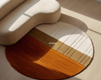 Peut inclure: Un tapis rond avec un design en blocs de couleurs dans les tons orange, beige et blanc. Le tapis a une surface texturée et est partiellement recouvert par un canapé beige incurvé. Le texte "ArtfulRugss" est visible sur le tapis.