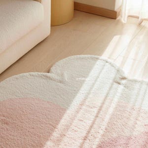 Peut inclure: Un tapis en forme de nuage avec une texture douce et moelleuse. Le tapis pr&eacute;sente un d&eacute;grad&eacute; de couleurs, passant d'un rose clair en bas &agrave; un blanc cr&egrave;me en haut. Le tapis est pos&eacute; sur un plancher en bois clair.