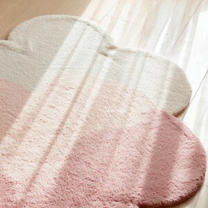 Peut inclure: Un tapis en forme de nuage, doux, dans des tons cr&egrave;me et rose. Le tapis a une surface moelleuse et textur&eacute;e et est pos&eacute; sur un sol en bois. Le design est simple et moderne, adapt&eacute; &agrave; une chambre ou une cr&egrave;che.