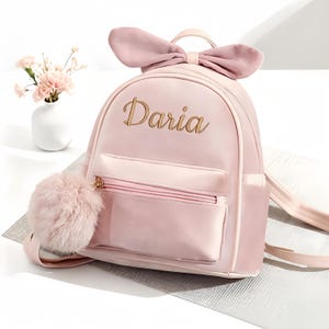 May include: A pale pink backpack with a large bow detail and a fluffy pom-pom. The name "Daria" is written in gold script. The bag has a front pocket and adjustable straps. A small vase of pink flowers is in the background.