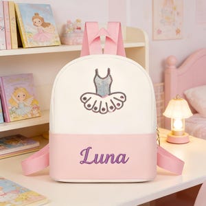 Puede incluir: Una mochila blanca y rosa con un diseño de tutú de bailarina. La mochila tiene correas rosas y el nombre "Luna" bordado en morado. La mochila está sobre un escritorio blanco con una lámpara y libros en el fondo.