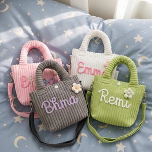 Puede incluir: Cuatro bolsos tote de pana en rosa, blanco, gris y verde, cada uno con un asa y una correa para el hombro a juego. Las bolsas tienen nombres bordados y un adorno floral. Las bolsas están sobre una superficie azul estampada.