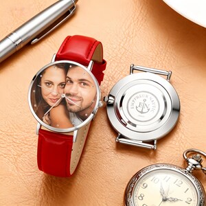 May include: A red leather strap watch with a silver case and a custom photo on the face. The watch face features a photo of a couple. The watch is next to a silver pen and a pocket watch.