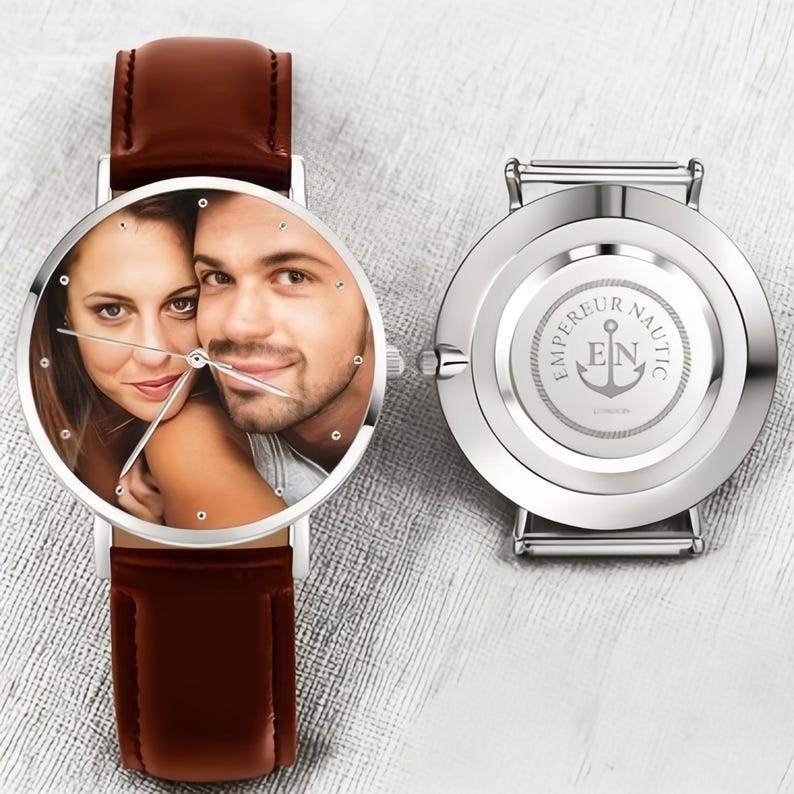 May include: A silver wristwatch with a brown leather band. The watch face displays a color photograph of a couple. The back of the watch has the words "EMPEREUR NAUTIC" and an anchor logo.