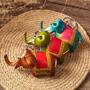 Pode incluir: Três porta-chaves em forma de elefante feitos de couro, cada um com um esquema de cores distinto em castanho, verde ou azul com painéis contrastantes em vermelho ou rosa e padrões decorativos amarelos nas bordas, presos a argolas de metal prateado.